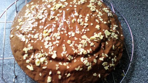 Ein Nusskuchen mit einer glänzenden, schokoladigen Glasur und gehobelten Nüssen auf der Oberfläche steht auf einem Gitter.