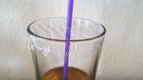 Ein Glas mit einem Getränk kann man ganz einfach mithilfe einer Frischhaltefolie vor Insekten und Schmutz schützen. Ein Glas mit Getränk wird durch eine Plastikfolie vor Schmutz und Insekten geschützt, während ein lila Strohhalm darin steckt.