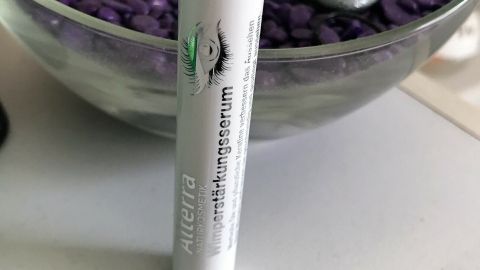 Ein Wimpernstärkungsserum steht auf einem Tisch, umgeben von lila Dekorsteinen und silbernen Dekorationen.
