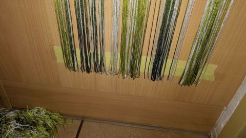 Mithilfe eines Klebebandstreifens lässt sich ein Fadenvorhang einfacher gleichmäßig kürzen. Ein farbiger Fadenvorhang hängt vor einer Tür, während einige Fäden am Boden liegen, was auf einen Kürzungsprozess hinweist.