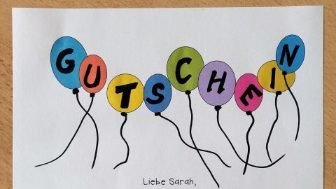 Bunte Luftballons mit dem Schriftzug „Gutschein“ ziehen Aufmerksamkeit auf einen kreativ gestalteten Gutschein für Kinder. Text: "Löse diesen Gutschein ein an einem Tag deiner Wahl, um sein neues Produkt selbst zu gestalten!"