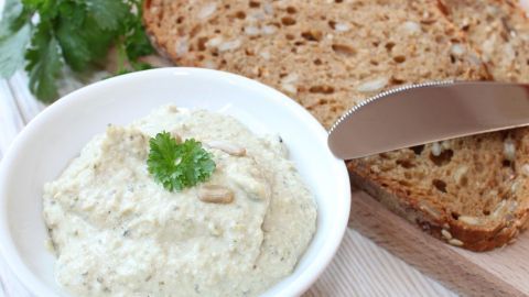 Kräuterdip mit Bockshornklee liegt in einer weißen Schüssel neben einer Brotscheibe auf einem Holzbrett.