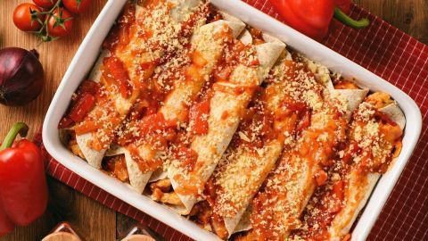 Eine Auflaufform mit sorgfältig gerollten, mit Sauce und Käse überbackenen Chicken Enchiladas steht auf einem rustikalen Tisch.
