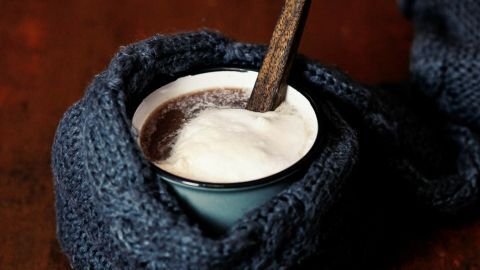 Eine Tasse heiße Schokolade mit einer Holzlöffel sitzt in einem blauen Schal auf einem dunklen Holztisch.
