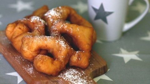 Auf einem Holzbrett liegen mehrere frisch ausgebackene Kreppel, bestäubt mit Puderzucker, neben einer Tasse auf einem Sterne-Tisch.