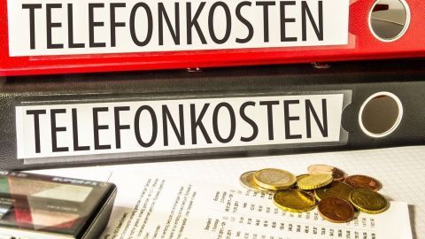 Zwei Aktenordner mit der Aufschrift „TELEFONKOSTEN“ stehen auf einem Tisch, umgeben von Münzen und einem Taschenrechner.