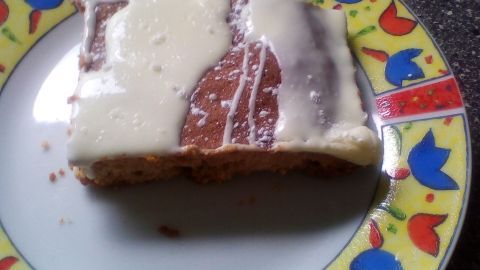 Ein Stück Tassenkuchen liegt auf einem bunten Teller, mit hellem Guss überzogen, vor einer dunklen Arbeitsplatte.