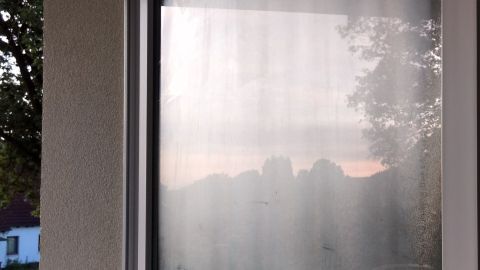 Eine beschlagene Fensterscheibe zeigt den Innenraum, während draußen Bäume und ein schwacher Sonnenuntergang sichtbar sind.