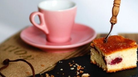 Ein Pflaumenkuchenstück liegt auf einem rustikalen Untergrund neben einer rosa Tasse auf einer Untertasse.