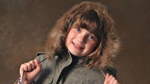 Ein Mädchen trägt eine warme Jacke mit Pelzkragen und lächelt fröhlich, während sie die Jacke im Studio präsentiert.