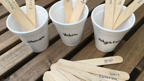 Drei Becher auf einem Tisch, gefüllt mit Holzstäbchen, die Wörter wie "Verb", "Substantiv" und "Adjektiv" beschreiben.