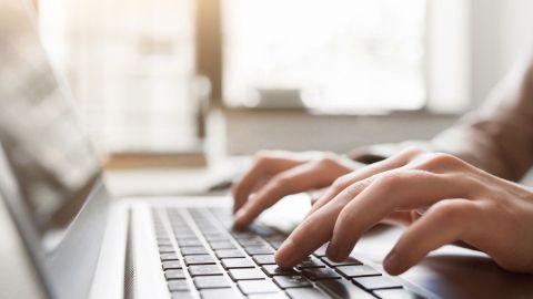 Eine Hand tippt auf einer Laptop-Tastatur in einem hellen Raum, um Texte zu schreiben und den Schreibstil zu verbessern.