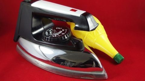 Ein Bügeleisen steht neben einer Zitronenflasche, bereit, mit Zitronensaft gereinigt zu werden, vor rotem Hintergrund.