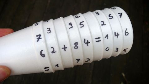 Ein Stapel weißer Becher mit handschriftlichen Zahlen und Matheaufgaben wird in einer Hand gehalten, um Mathe für coole Kids zu veranschaulichen.