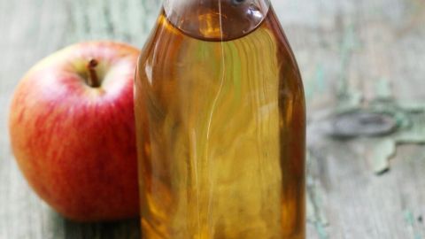 Ein Glas mit Essig steht neben einem roten Apfel auf einer rustikalen Holzoberfläche. Essig kann bei Stielwarzen helfen.