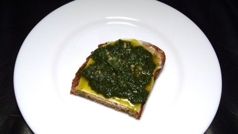 Ein Stück Brot liegt auf einem weißen Teller, das mit grünem Pesto als Brotaufstrich bestrichen ist.