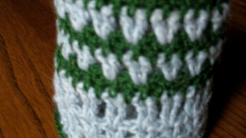 Ein gehäkelter Becherhalter aus Wollresten in Grün und Weiß steht auf einem Holztisch und präsentiert ein dekoratives Muster.