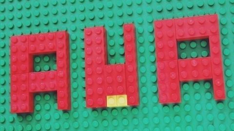 Rote Legosteine formen das Wort "AUA" auf einer grünen Unterlage, was auf Rückenschmerzen bei Verspannungen hinweist.