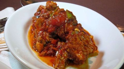 Ein geschmorter Eintopf liegt in einer weißen Schüssel, umgeben von Tomatensauce und Gemüse, typisch für Coda alla vaccinara.