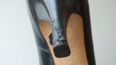 Ein schwarzer High Heel steht im Fokus, der an der Spitze beschädigt ist. Die Umgebung ist neutral und hell.