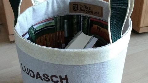 Ein runder Bücherkorb aus SnapPap und Stoff steht auf einem hellen Fußboden, gefüllt mit Bücher und hat grüne Tragegriffe.