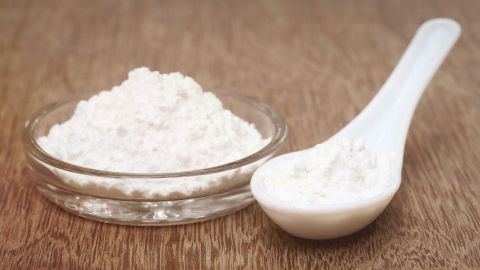Ein Schälchen mit feinem, weißem Puder steht neben einem Löffel, der auf einer hölzernen Oberfläche liegt. Sanft reinigen.
