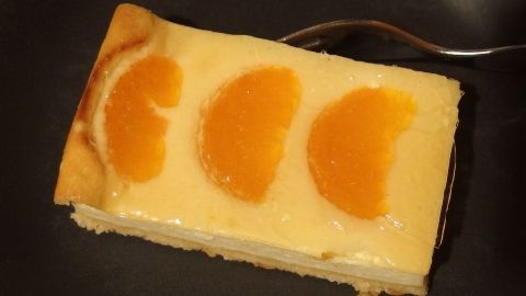 Ein Stück Käsekuchen mit orangefarbenen Fruchtscheiben liegt auf einem dunklen Teller, bereit zum Genießen.