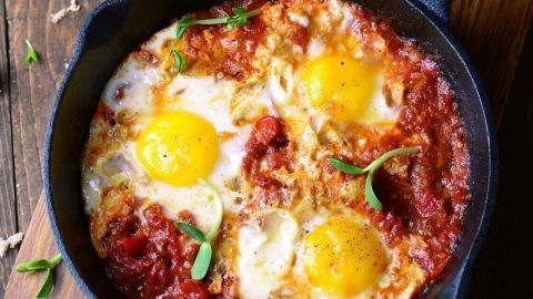 Eine Pfanne mit Shakshuka, vier Spiegeleiern in Tomatensauce, steht auf einem Holzbrett, umgeben von frischen Zutaten.