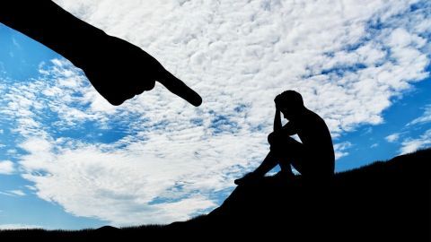 Eine Hand zeigt auf eine sitzende Person, die sich den Kopf hält, unter einem weiten, wolkigen Himmel.