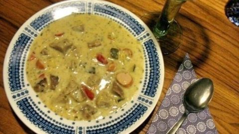 Eine bunte Brotsuppe mit Frischkäse steht in einer dekorativen Schale auf einem Holztisch, umgeben von Besteck und einem Getränk.