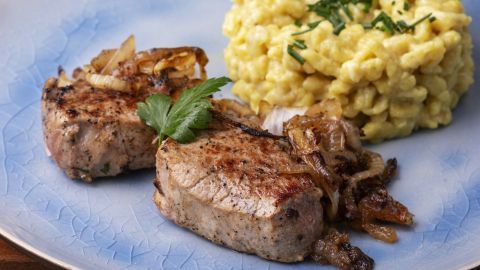 Saftige Colabratenstücke liegen mit Zwiebeln auf einem blauen Teller, begleitet von gelbem Risotto mit Schnittlauch.