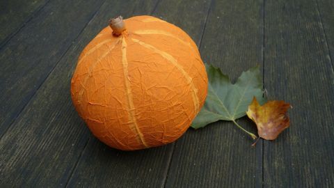 Ein Kürbis aus Pappmaché liegt auf einem Holzfußboden, umgeben von einigen farbigen Blättern.