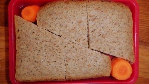 Eine rote Brotbox enthält unregelmäßig geschnittenes Vollkornbrot und orange Karottenstückchen in einer ungeordneten Anordnung.