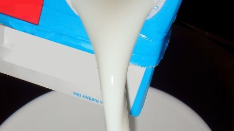 Ein Milchkarton wird über eine Tasse gehalten, während frische Milch langsam hineinfließt, ideal für Rezepte und Kochbuchideen.