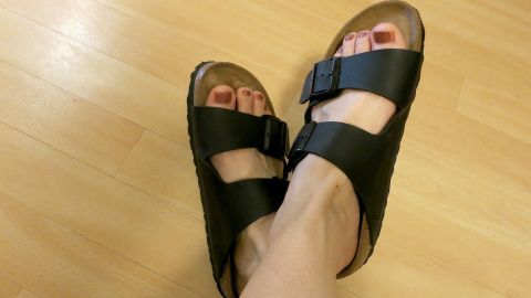 Sandalen am Fuß ruhen auf hellem Holzboden; schwarzes Leder mit Schnallen, entspanntes und komfortables Ambiente.
