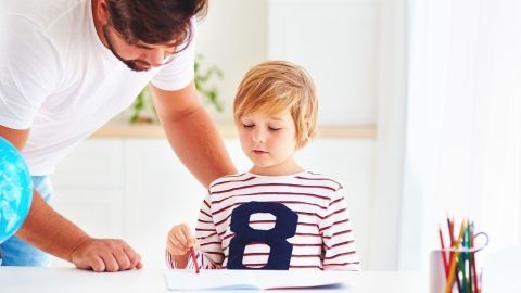 Ein Erzieher hilft einem Kind, während sie gemeinsam am Tisch arbeiten, umgeben von Materialien und einem hellen Raum.