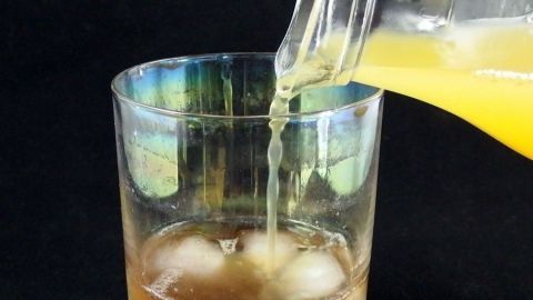 Ein Glas wird mit einer orangefarbenen Flüssigkeit gefüllt, während Eiswürfel im Glas liegen, vor dunklem Hintergrund.