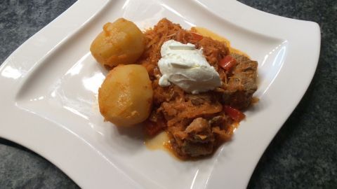 Auf einem weißen Teller liegt Szegediner Krautfleisch, umgeben von Kartoffeln und einem Klecks Sauerrahm.