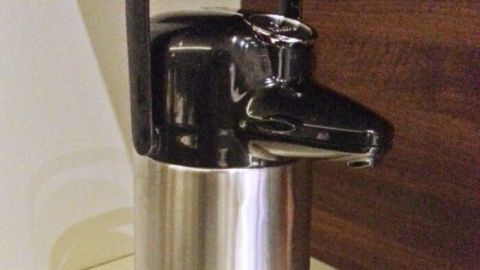 Eine glänzende Thermoskanne steht auf einer Oberfläche und scheint Kaffee zu enthalten, mit einem Ausgießer oben.