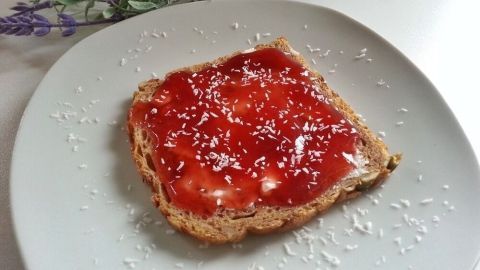 Ein Stück Brot liegt auf einem Teller und ist mit Erdbeer-Kokos-Marmelade und Kokosraspeln dekoriert.