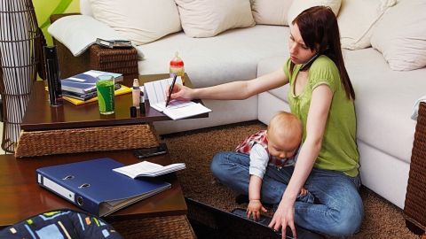 Eine Frau mit einem Baby auf dem Schoß multitaskt, indem sie am Laptop arbeitet und Notizen macht, während sie zu Hause sitzt.