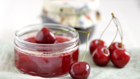 Ein Glas Marmelade steht auf einem Tisch, umgeben von frischen Kirschen, die appetitlich glänzen.