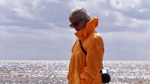 Eine Person in einem gelben Regenmantel geht am Strand entlang, während das Sonnenlicht auf das Wasser reflektiert.