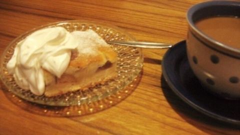 Ein Blechkuchen mit Sahne liegt auf einem gläsernen Teller neben einer Tasse Kaffee auf einem Holztisch.