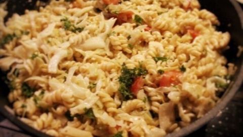 Eine Pfanne mit bunt gemischten Nudeln, Lachs und Spitzkohl wird in einer dunklen Küche serviert.