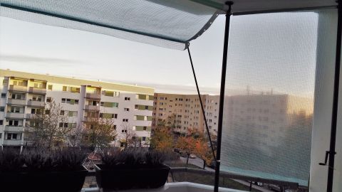 Eine Wintermarkise schützt den Balkon vor Nässe, während im Hintergrund Wohnblöcke und ein sanfter Sonnenuntergang sichtbar sind.