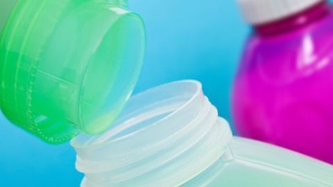 Eine grüne Weichspülerflasche wird über eine offene Flasche gegossen, während eine pinke Flasche im Hintergrund sichtbar ist.