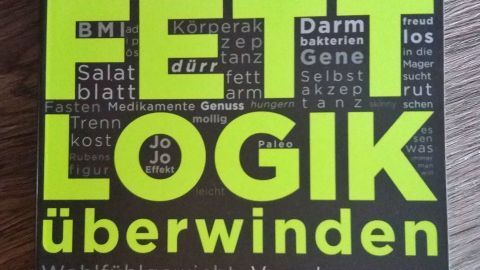 Ein schwarzes Buch mit dem Titel „FETTLOGIK überwinden“ von Dr. Nadja Hermann liegt auf einem Holzuntergrund.