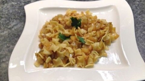 Krautfleckerl auf einem eleganten weißen Teller, gebraten mit Zwiebeln und mit Kräutern garniert, auf einer grauen Oberfläche.