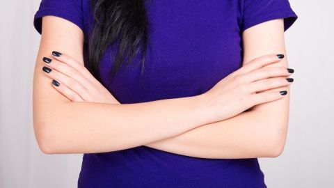 Eine Person mit schwarzen Nägeln steht mit verschränkten Armen und trägt ein lila T-Shirt, was eine defensive Körpersprache zeigt.